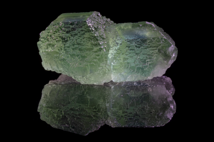 Horizontal full specimen view of two linked pale green etched fluorite cubes with textured surfaces and mirror reflection on black background — macro photography by Minerals Photography