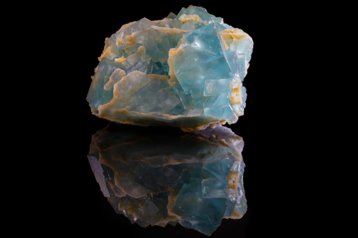 Full specimen view of aqua blue-green fluorite cubic crystals with cream-yellow gangue matrix and mirror reflection on black background — macro photography by Minerals Photography