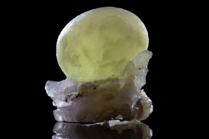 Single large yellow-green botryoidal fluorite sphere resting on white quartz crystal cluster with black mineral inclusions and mirror reflection on black background — macro photography by Minerals Photography