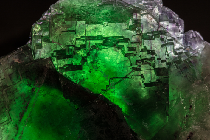 Extreme macro of etched fluorite crystal face showing luminous emerald green interior glowing through stepped dissolution terraces with violet color zoning at edges — macro photography by Minerals Photography