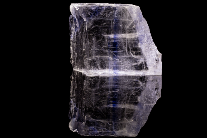 Full specimen view of blue-banded halite cubic crystal with deep blue horizontal zones visible through translucent body, mirror reflection on black background — macro photography by Minerals Photography