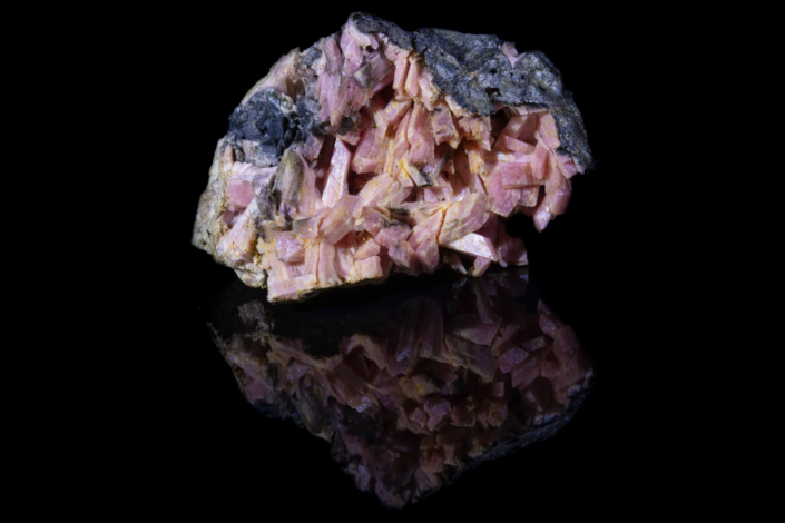 Full specimen view of pink-mauve tabular heulandite crystals on dark gray matrix with mirror reflection on black background — macro photography by Minerals Photography