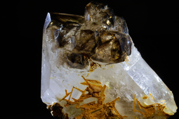 Close-up of smoky quartz cluster on colorless quartz showing dark secondary crystals and orange mineral inclusions at the base, macro photography by Minerals Photography