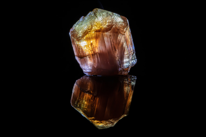 Scapolite crystal with amber and rose-violet color zoning under backlight reflected on black mirror surface, macro photography by Minerals Photography