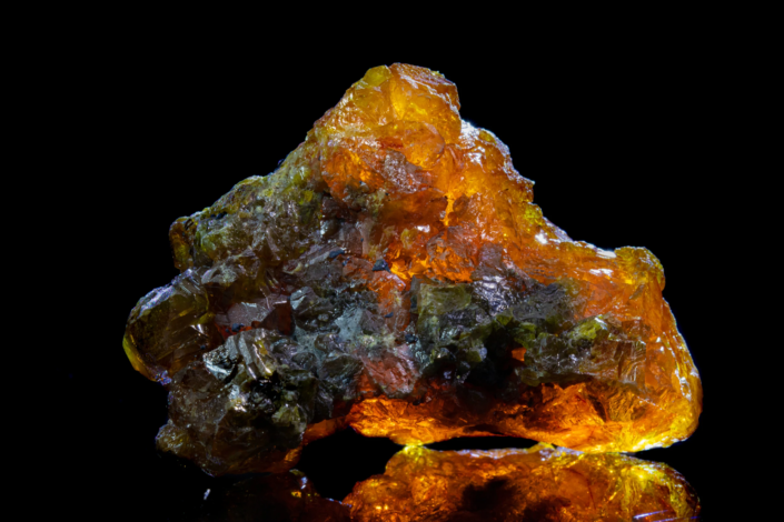 Sphalerite specimen glowing amber-orange under backlight with green and dark crystal zones reflected on black mirror surface, macro photography by Minerals Photography